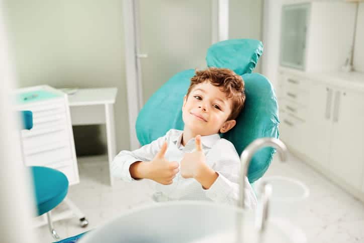 A child giving two thumbs up while sitting in a dental chair at a pediatric dentist's office. 
