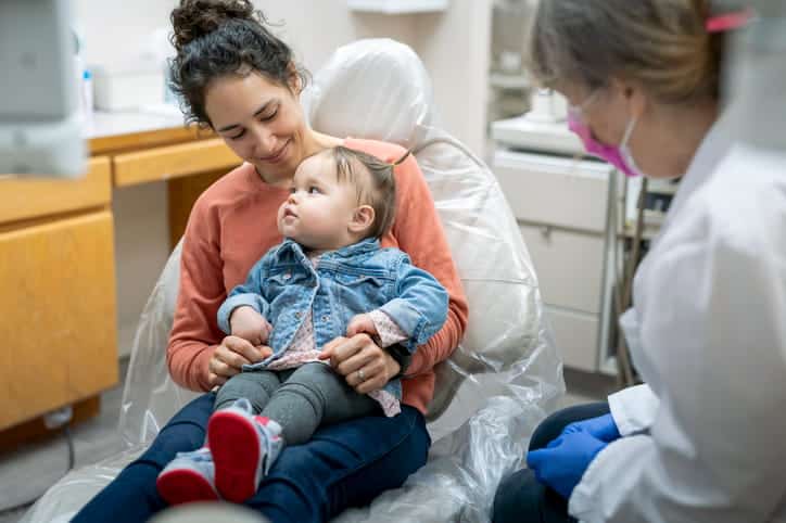 A child sitting on their mother's lap during their first dental visit at the pediatric dentist's office. 