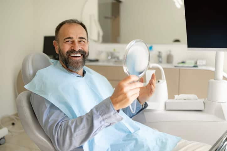 A man smiling while holding a mirror at the dentist's office. 