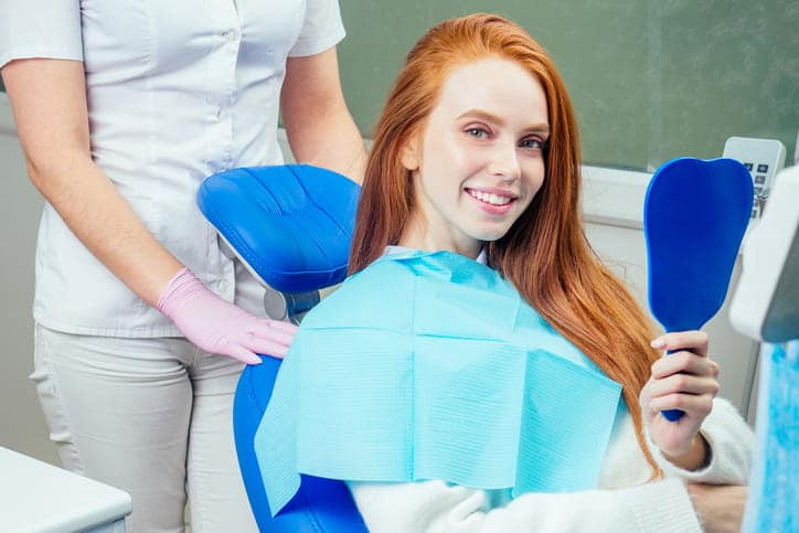 A woman smiling and holding a mirror at the dentist's office after finishing treatment for a gum lift. 