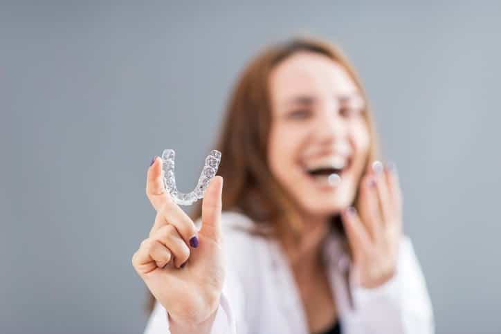 A woman is out of focus as she laughs, holding a clear aligner that is in focus.