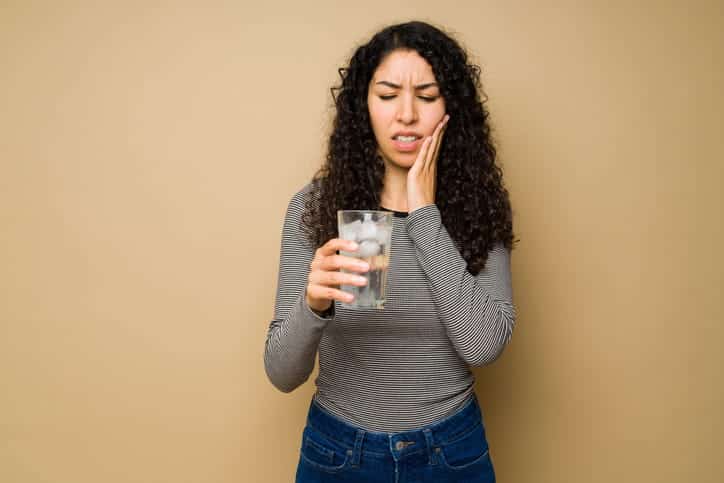 A woman holding her face in pain while holding a glass of water because of wisdom teeth pain.