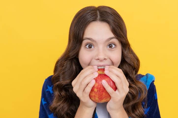 A young girl biting into an apple with a yellow background.