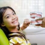Focus is on a little girl smiling at the camera as she points to the 3D model of teeth with metal braces that a dentist is handing her.