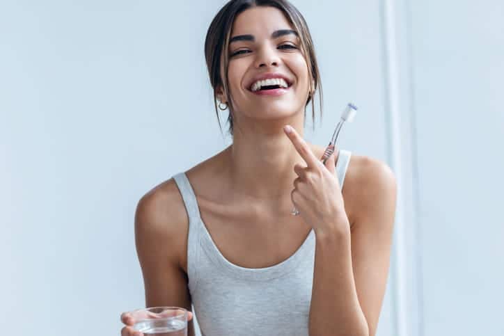 A woman holding a toothbrush and a glass of water pointing at her smile. 