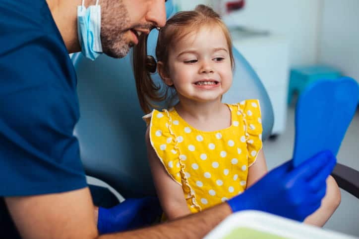 A little girl smiles into the mirror her pediatric dentist is holding.