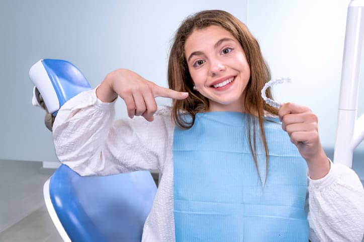 A teenage girl is at the dentist's office smiling as she points at her Invisalign aligner.