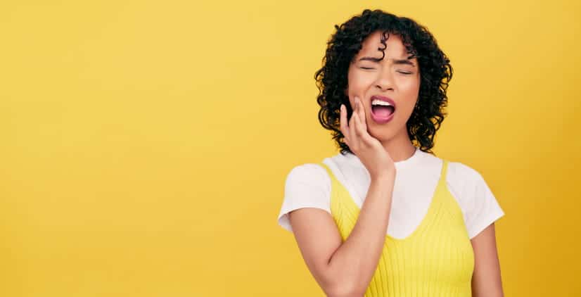 A woman in front of a yellow background, holding her face in pain due to tooth sensitivity.