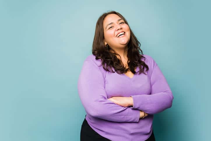 A woman smiling with her arms crossed in front of a teal background.