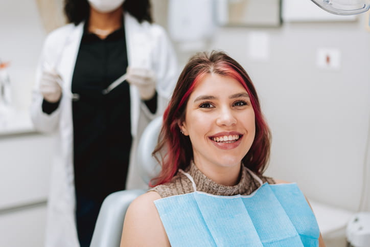 A woman smiling while sitting in a dental chair. The dentist is blurred out in the background.